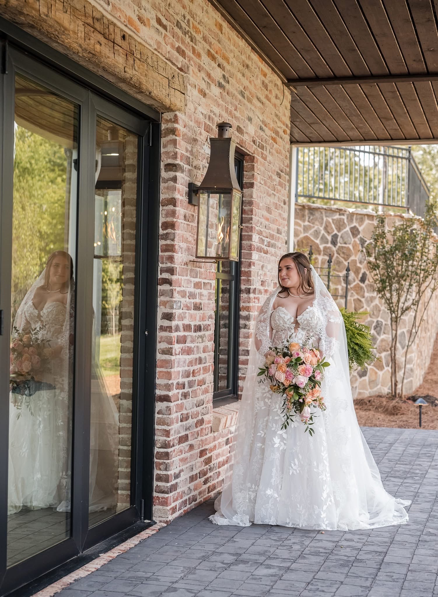 Bride in lace gown standing by the brick wall with her reflection in the glass door