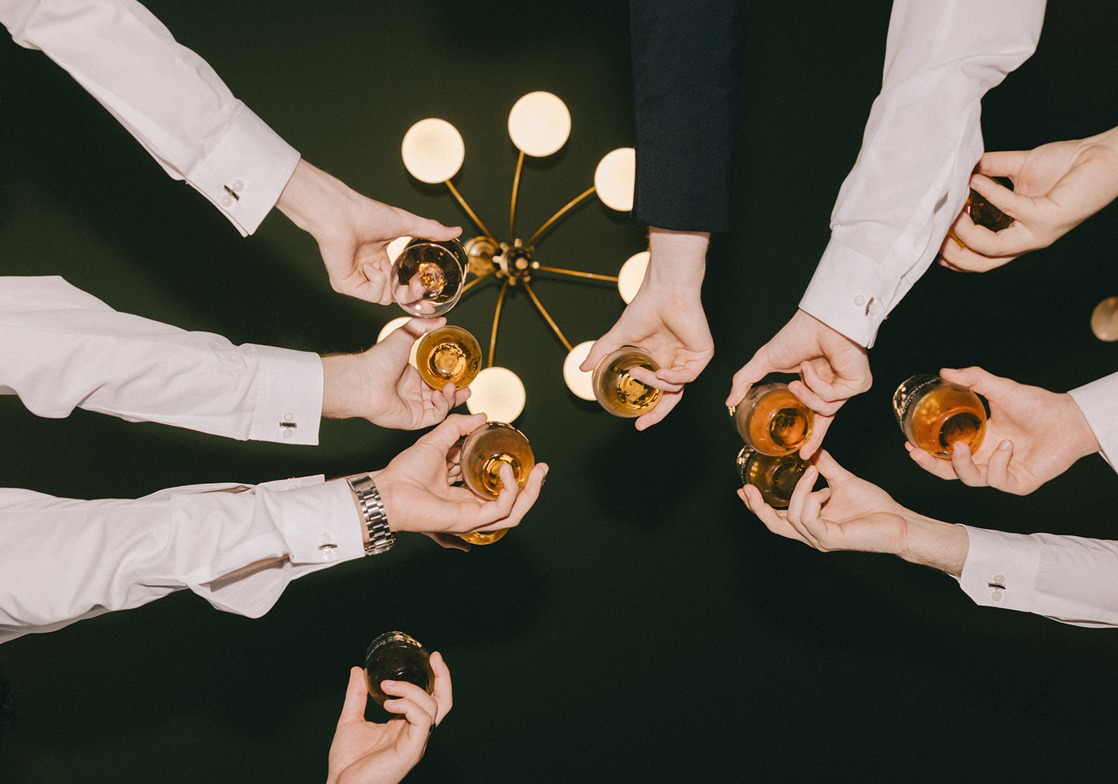 Overhead shot of groomsmen toasting with whiskey glasses under a gold chandelier